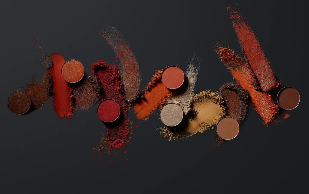 Eye shadow palettes and loose pigment in warm brown and orange tones arranged on a dark grey surface, high-angle studio shot.