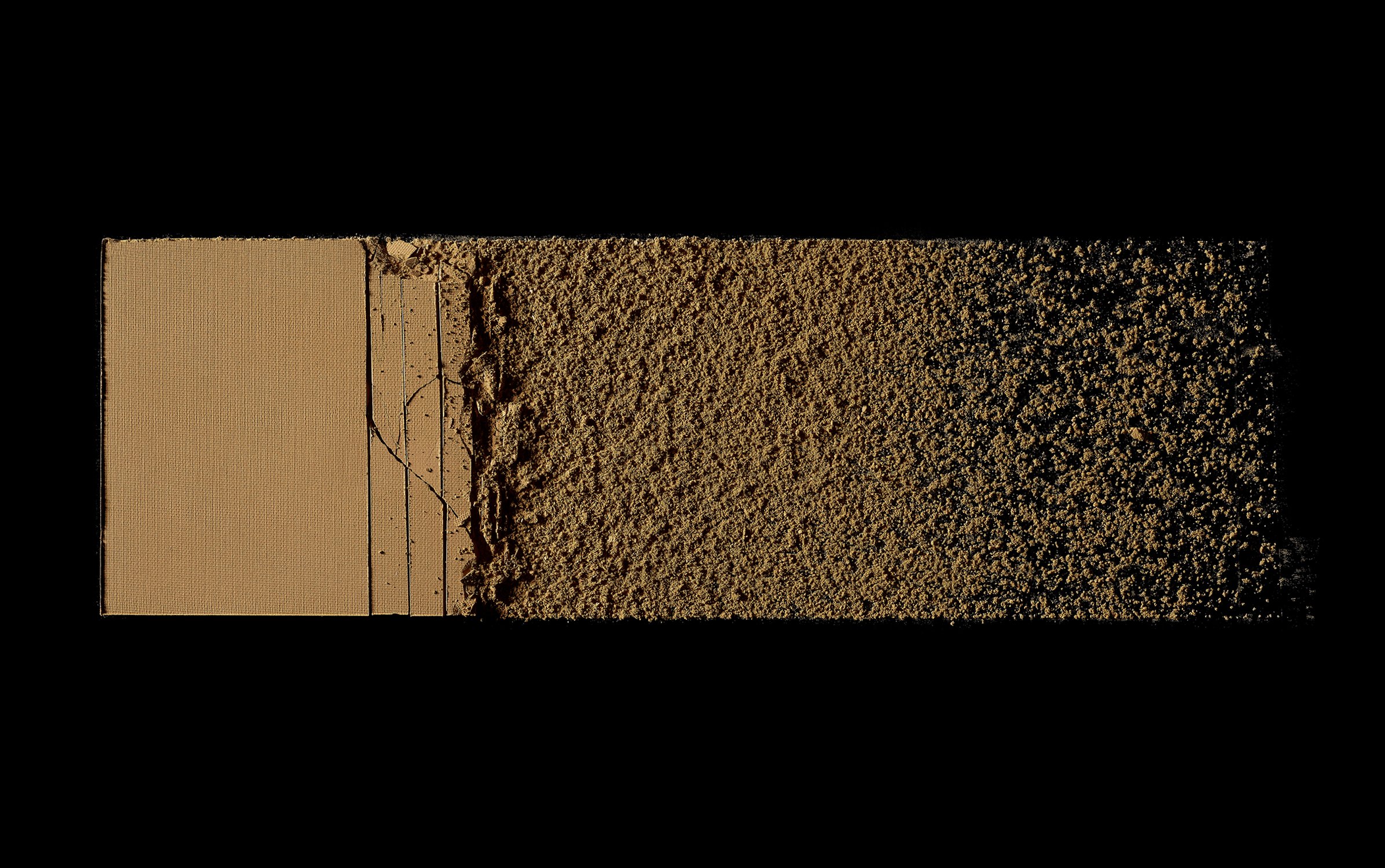 Close-up of a tan-colored pressed powder makeup palette, transitioning from solid to crumbled texture against a stark black background.