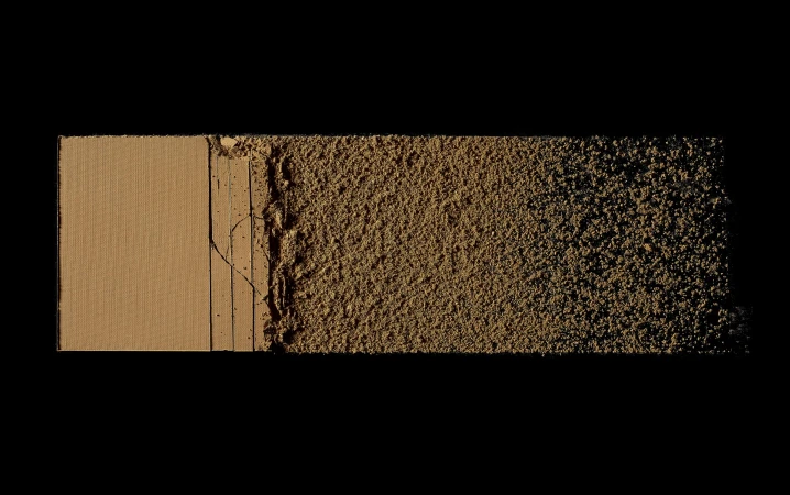 Close-up of a tan-colored pressed powder makeup palette, transitioning from solid to crumbled texture against a stark black background.