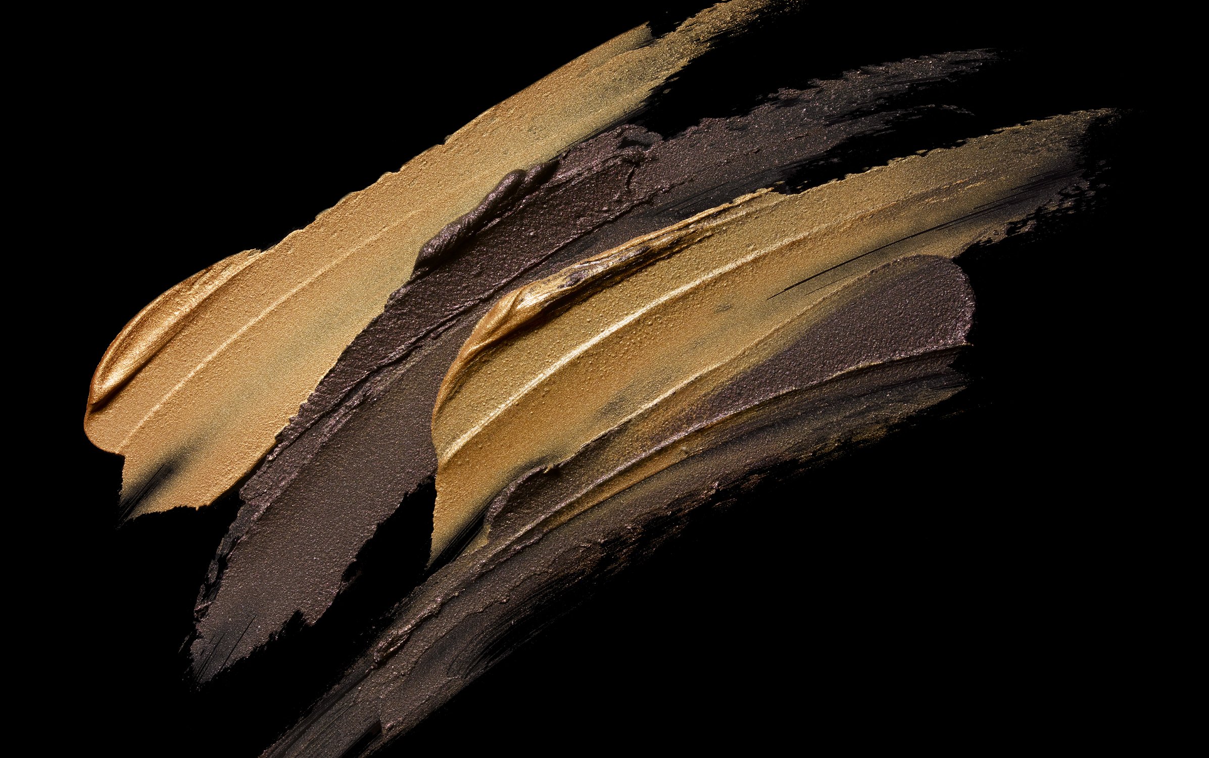 Close-up of gold and brown cosmetic product smears on a black background creating an abstract textural pattern, shot in studio.