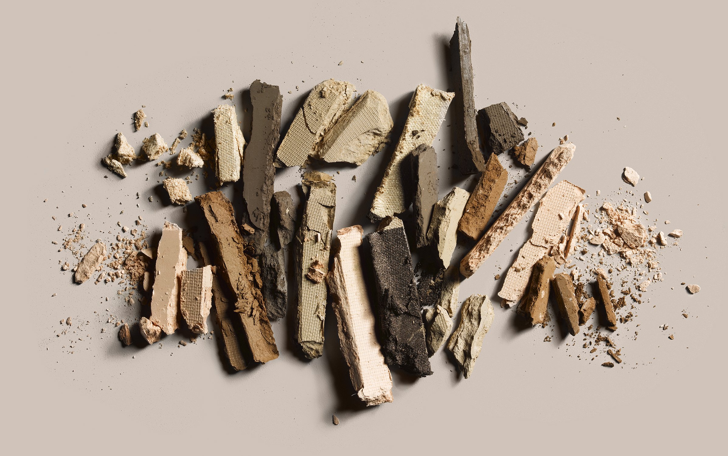 Crushed eyeshadow powder in neutral tones, including beige, brown, and black, scattered on a light background creating an abstract pattern.