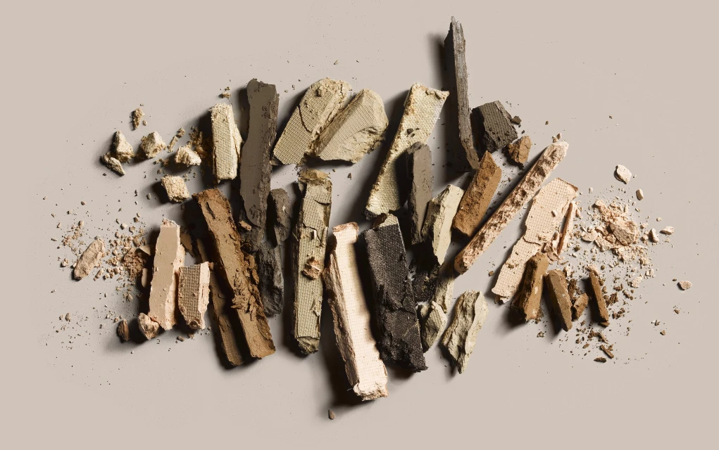 Crushed eyeshadow powder in neutral tones, including beige, brown, and black, scattered on a light background creating an abstract pattern.