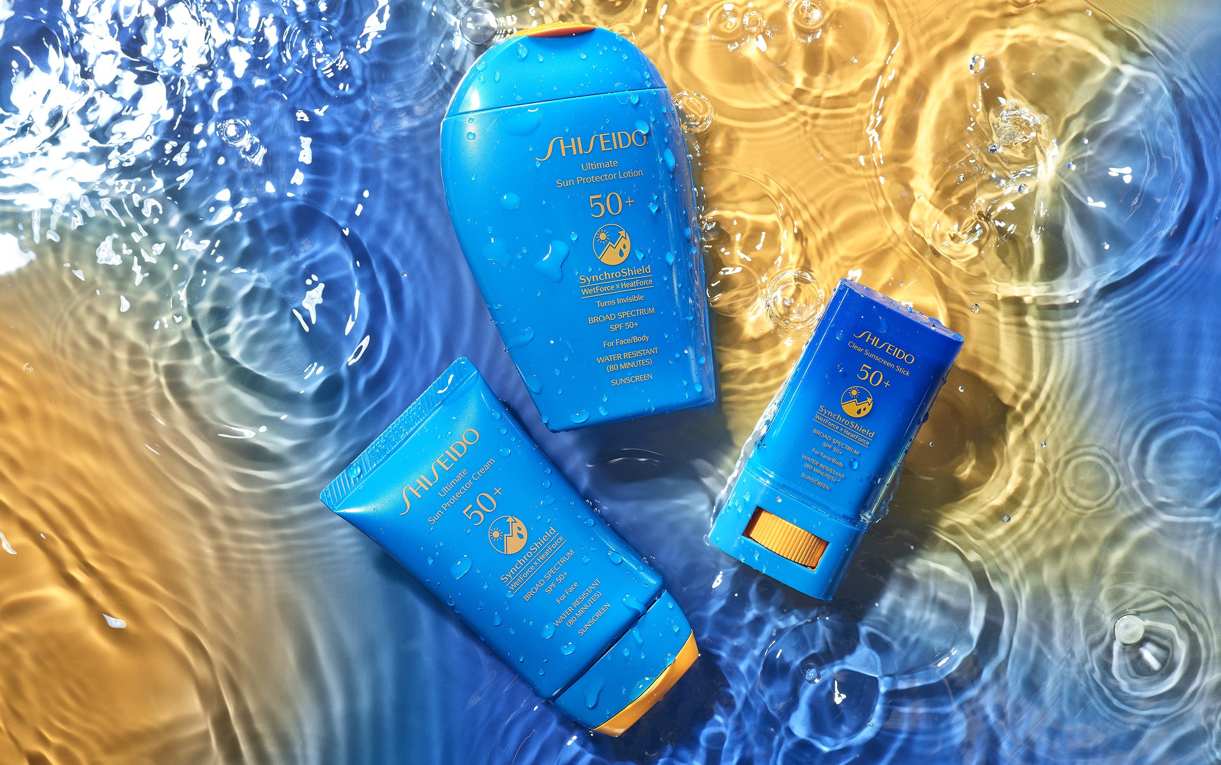 Three blue Shiseido sunscreen bottles, including lotion and stick, are arranged in shallow water with rippling blue and gold reflections.