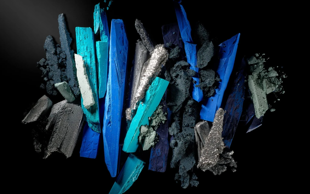 Abstract beauty shot featuring crushed eyeshadow in shades of blue, teal, gray, and silver, set against a stark black background.