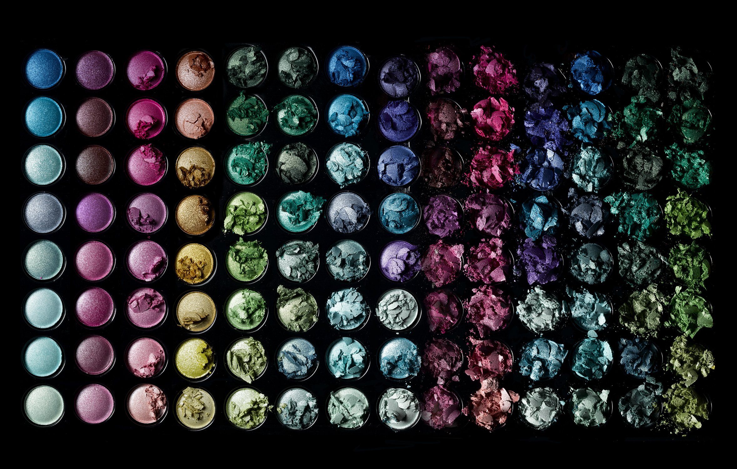 Overhead shot of a vibrant eyeshadow palette showcasing an array of colors including blues, greens, pinks, purples, and metallics, isolated on black.