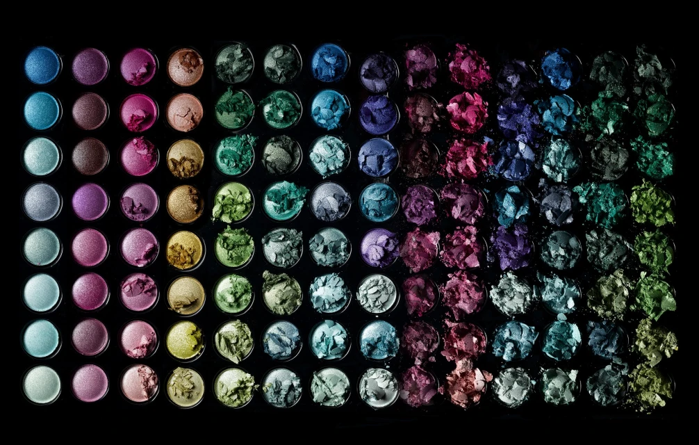 Overhead shot of a vibrant eyeshadow palette showcasing an array of colors including blues, greens, pinks, purples, and metallics, isolated on black.