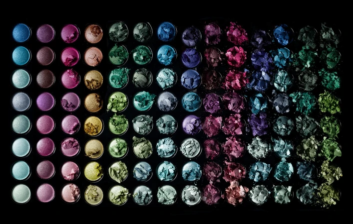 Overhead shot of a vibrant eyeshadow palette showcasing an array of colors including blues, greens, pinks, purples, and metallics, isolated on black.
