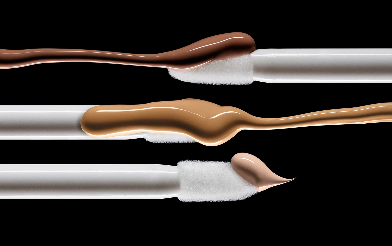 Three white cosmetic applicator wands displaying smears of liquid foundation in varying shades against black