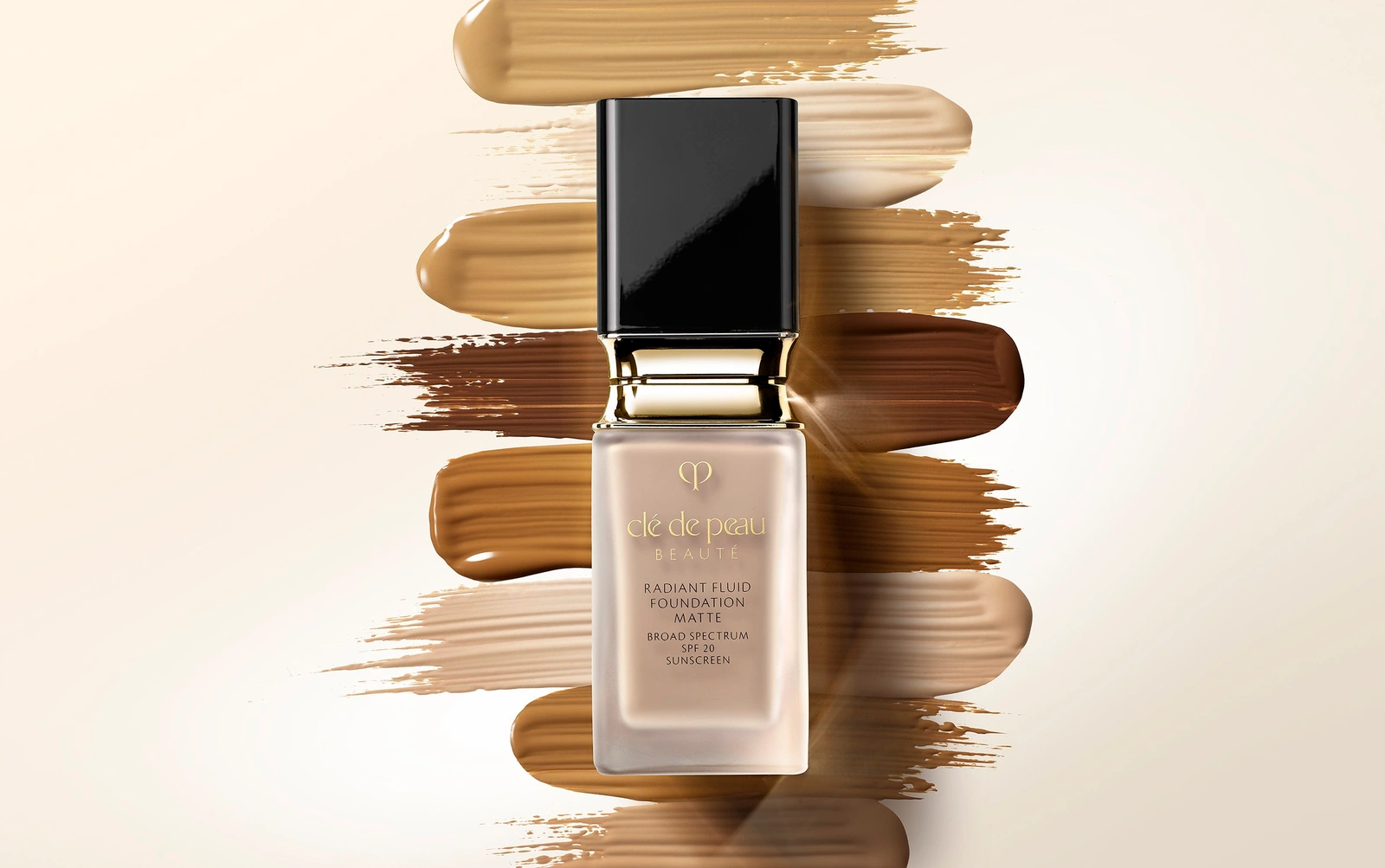 A rectangular glass bottle of liquid foundation with a black cap centered over horizontal brush strokes of various foundation shades on white.