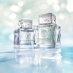 Two glass perfume bottles with water droplets on a reflective surface against a soft blue and white bokeh background.