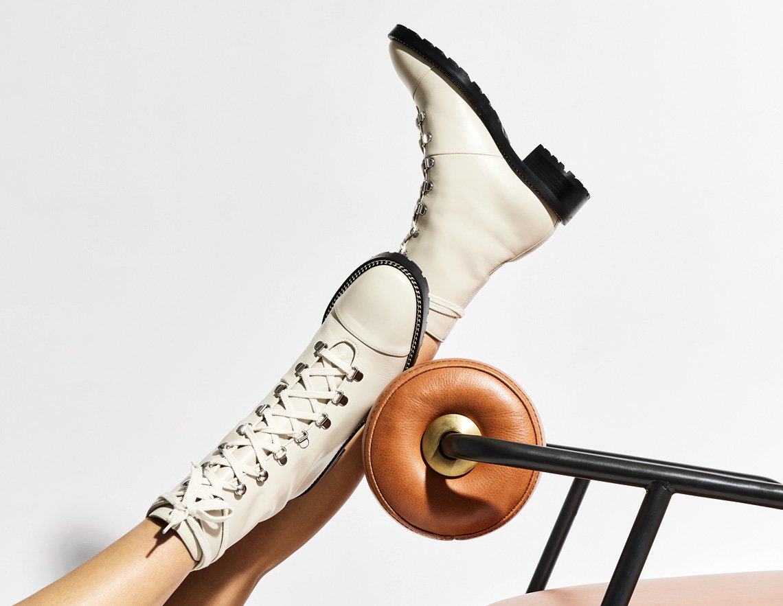 Fashion still life: A single leg is raised displaying a white leather, lace-up boot with a low black heel, and a brown and black chair.
