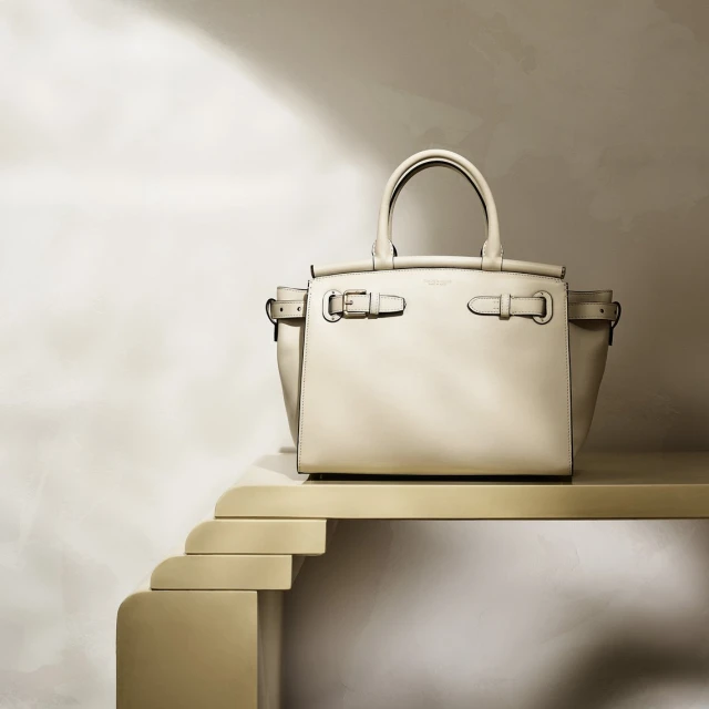 Beige leather handbag with top handles and silver hardware, placed on a golden tiered display against a light beige background.