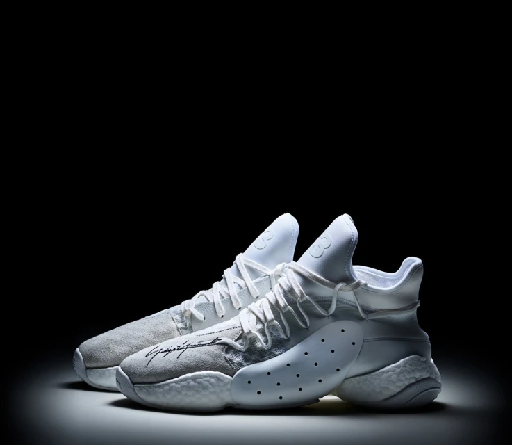 Two white Y-3 sneakers are showcased against a black backdrop, highlighted by a central spotlight that accentuates their design and features.