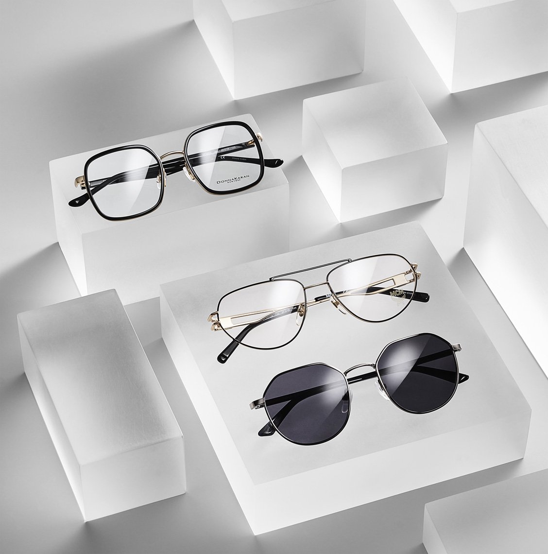 Studio shot of three pairs of eyeglasses with black, gold, and silver frames arranged on translucent white blocks against a white backdrop.