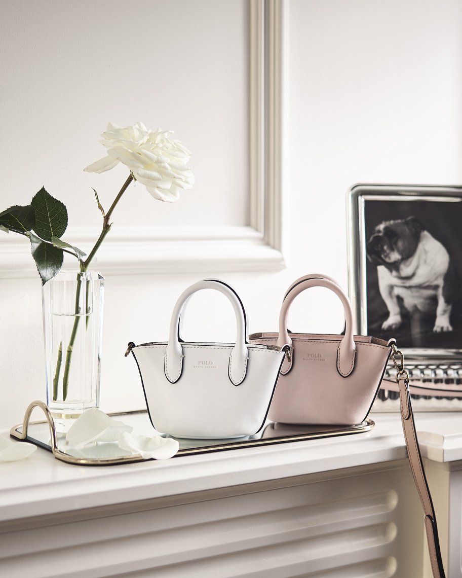 Two miniature leather tote bags, one white and one light pink, sit on a mirrored tray next to a white rose in a glass vase.