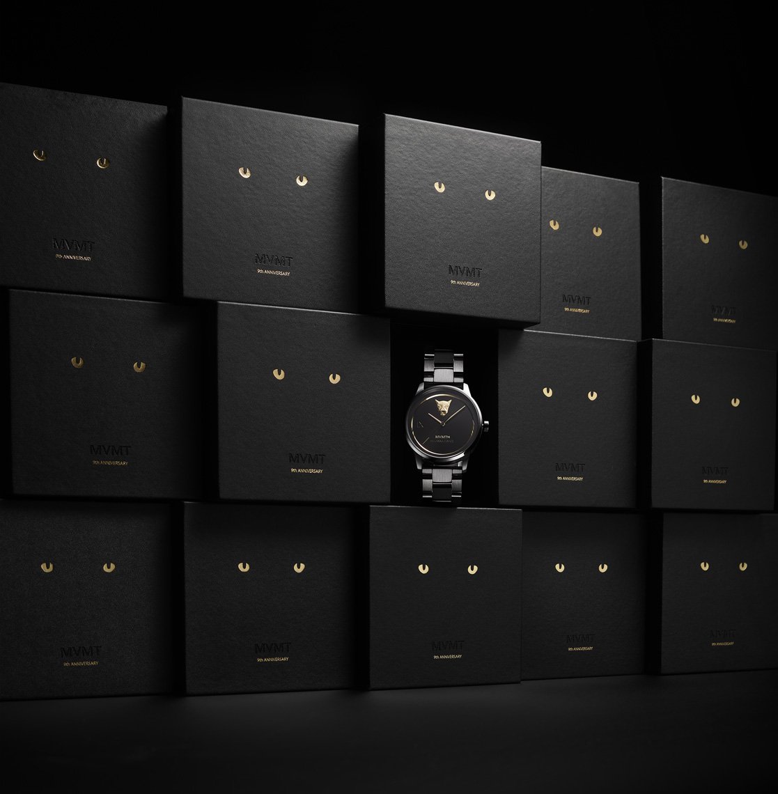 Wristwatch with a metal band displayed on a black surface surrounded by multiple black boxes with stylized gold cat-eye graphics.