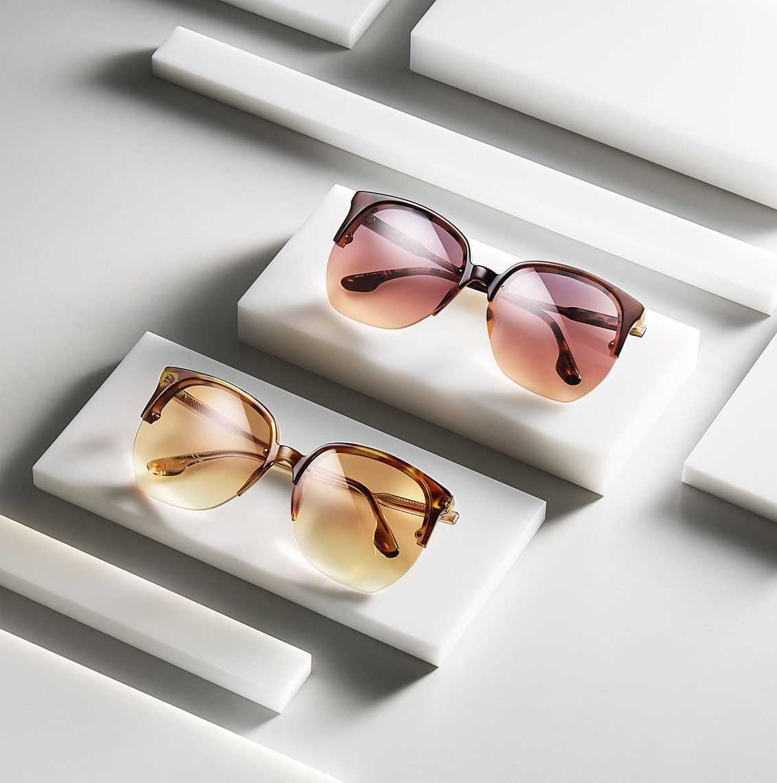 Two pairs of sunglasses are displayed on white blocks against a white background in a high-angle shot with diffused lighting.