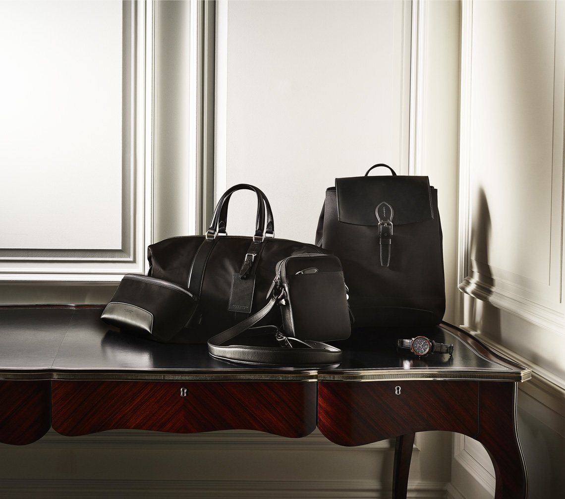 Luxury black leather duffel bag, backpack, and accessories arranged on a dark wood table against an elegant wall with panel molding.