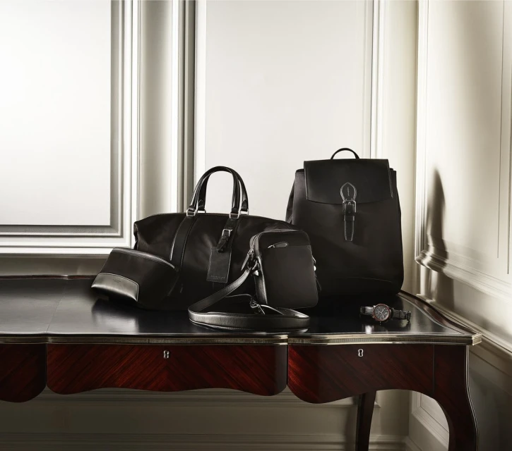 Luxury black leather duffel bag, backpack, and accessories arranged on a dark wood table against an elegant wall with panel molding.