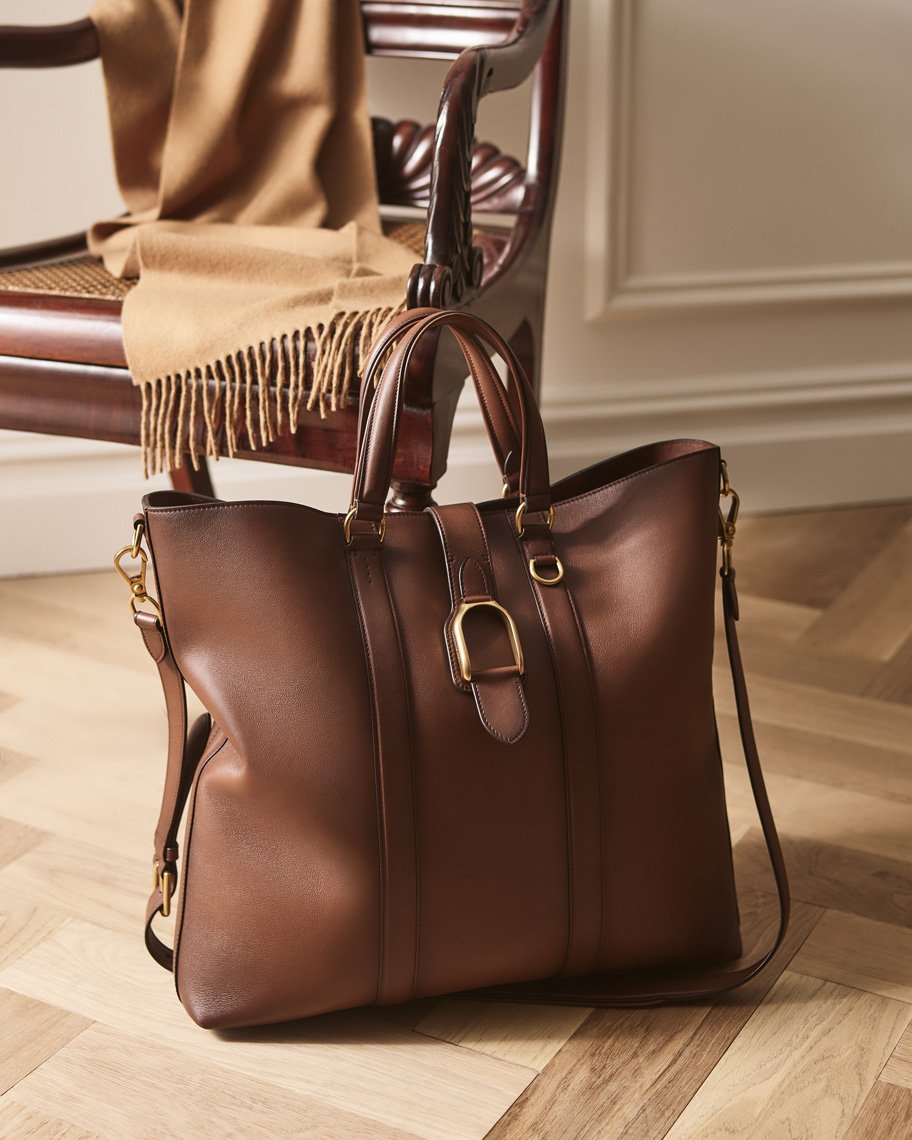 Brown leather tote bag with a gold buckle detail, placed on a wooden floor with a draped beige scarf on a chair in the background.