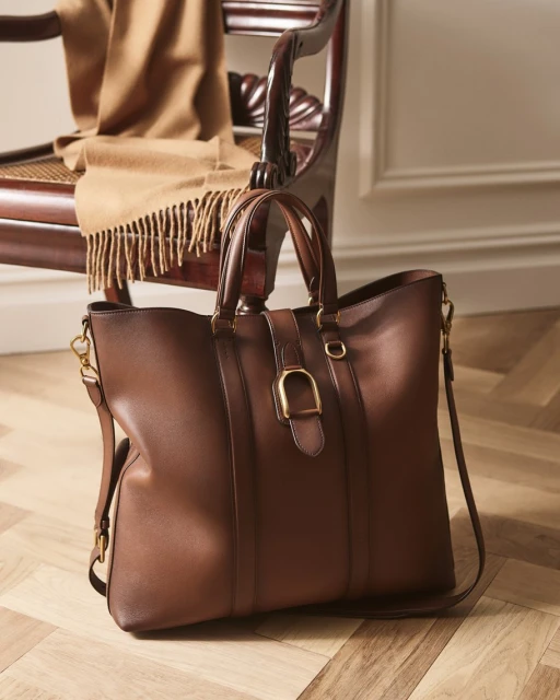 Brown leather tote bag with a gold buckle detail, placed on a wooden floor with a draped beige scarf on a chair in the background.