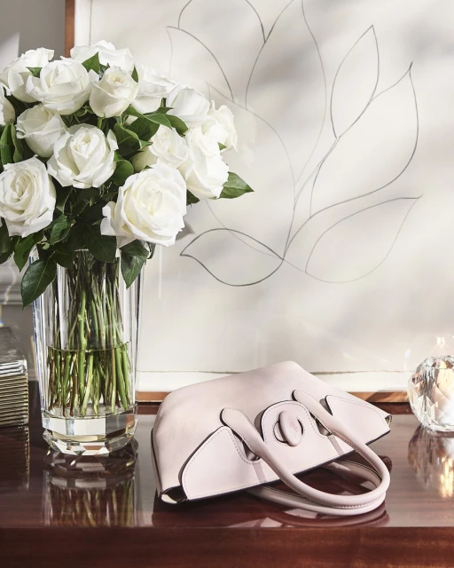 Elegant scene featuring a light pink purse on a dark wood table, complemented by white roses in a crystal vase and floral art.