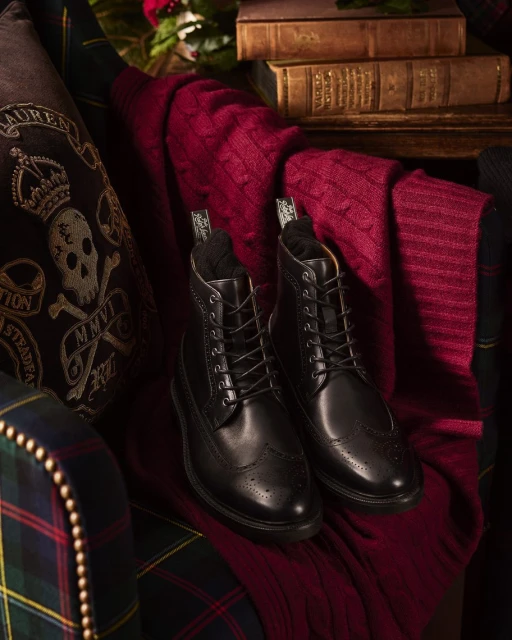 Still life of black leather wingtip boots with a red patterned scarf, books, and a pillow with a skull on a chair.
