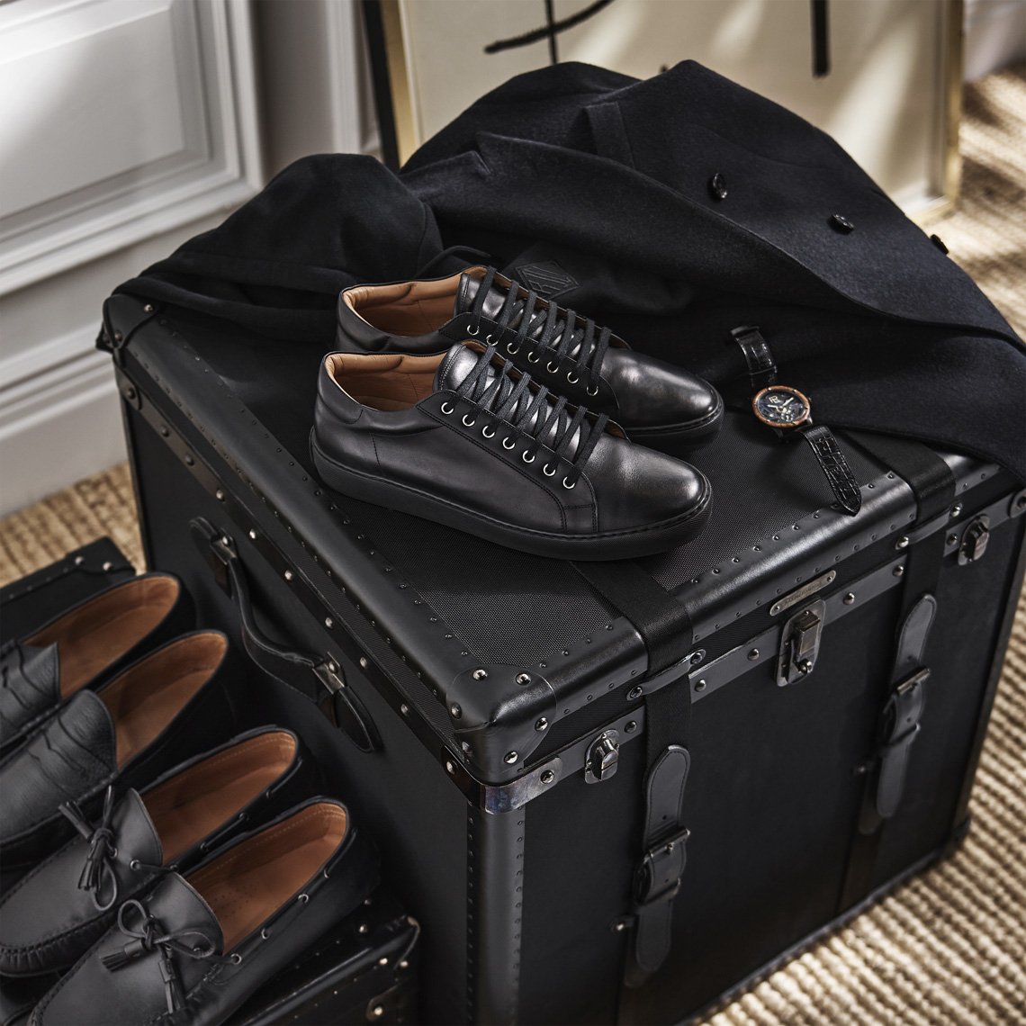 Elegant black leather shoes and a watch displayed on a vintage trunk, complemented by a stylish black coat. Fashionable and upscale.