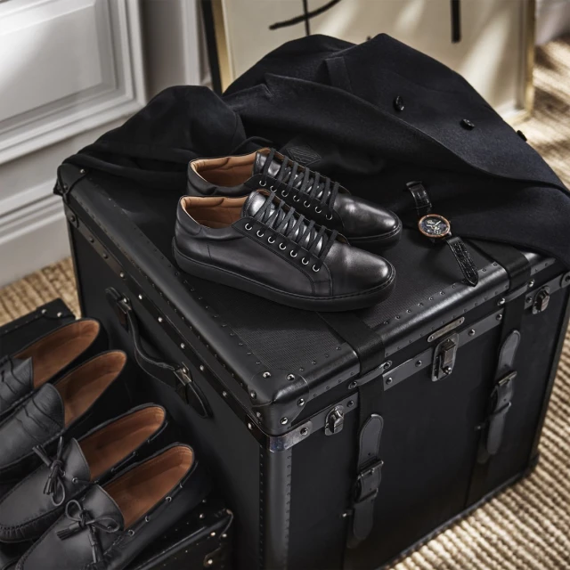 Elegant black leather shoes and a watch displayed on a vintage trunk, complemented by a stylish black coat. Fashionable and upscale.