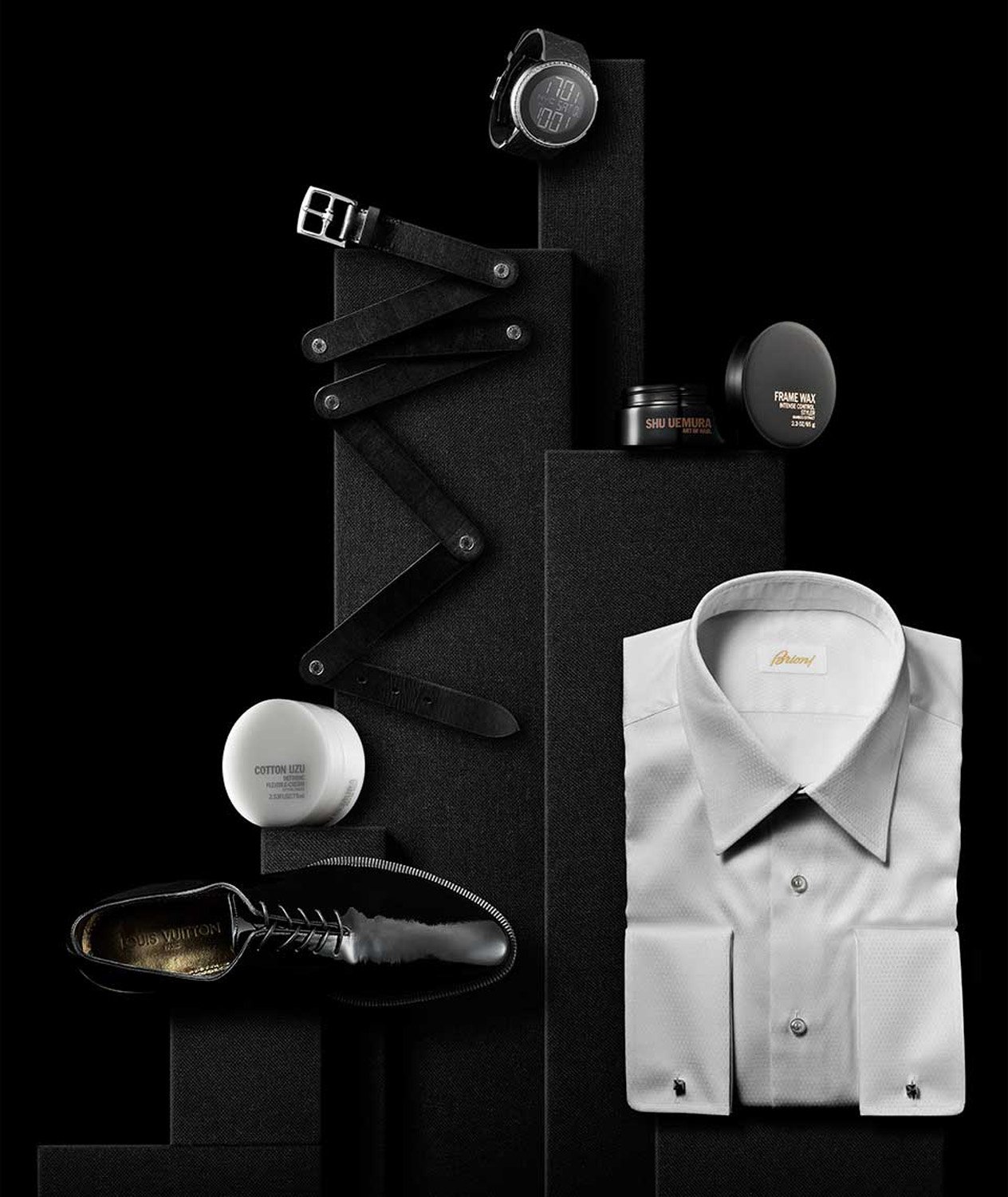 Elevated product still life in black and white featuring a dress shirt, belt, watch, skincare, and dress shoe, presented on blocks.