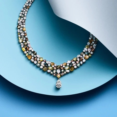 Elegant multi-colored gemstone necklace displayed on a light blue surface with a curved white paper accent in a studio setting.