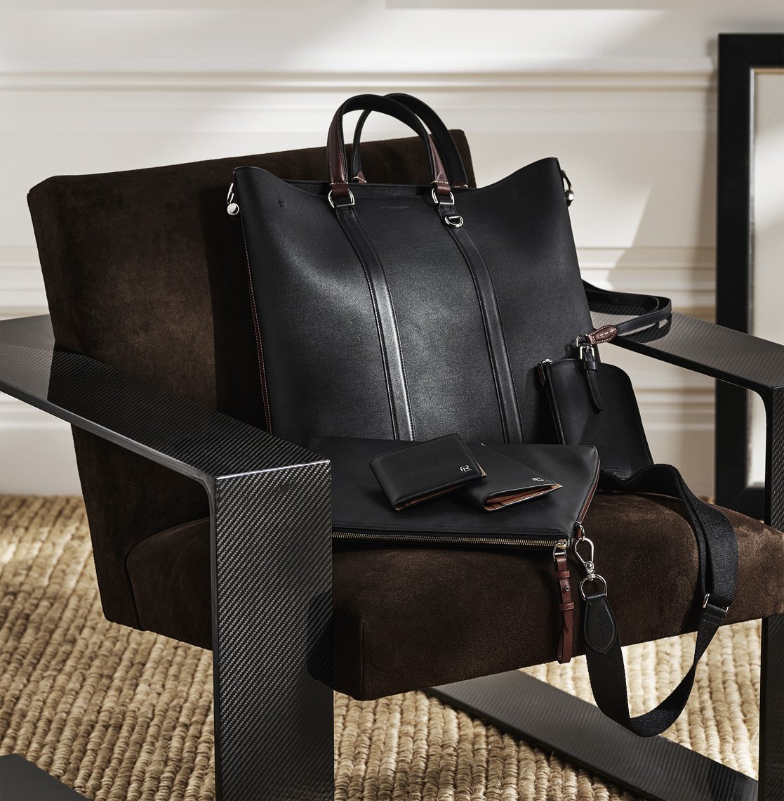 Still life of a black leather tote bag, wallet, and pouch arranged on a modern brown suede chair with carbon fiber accents.