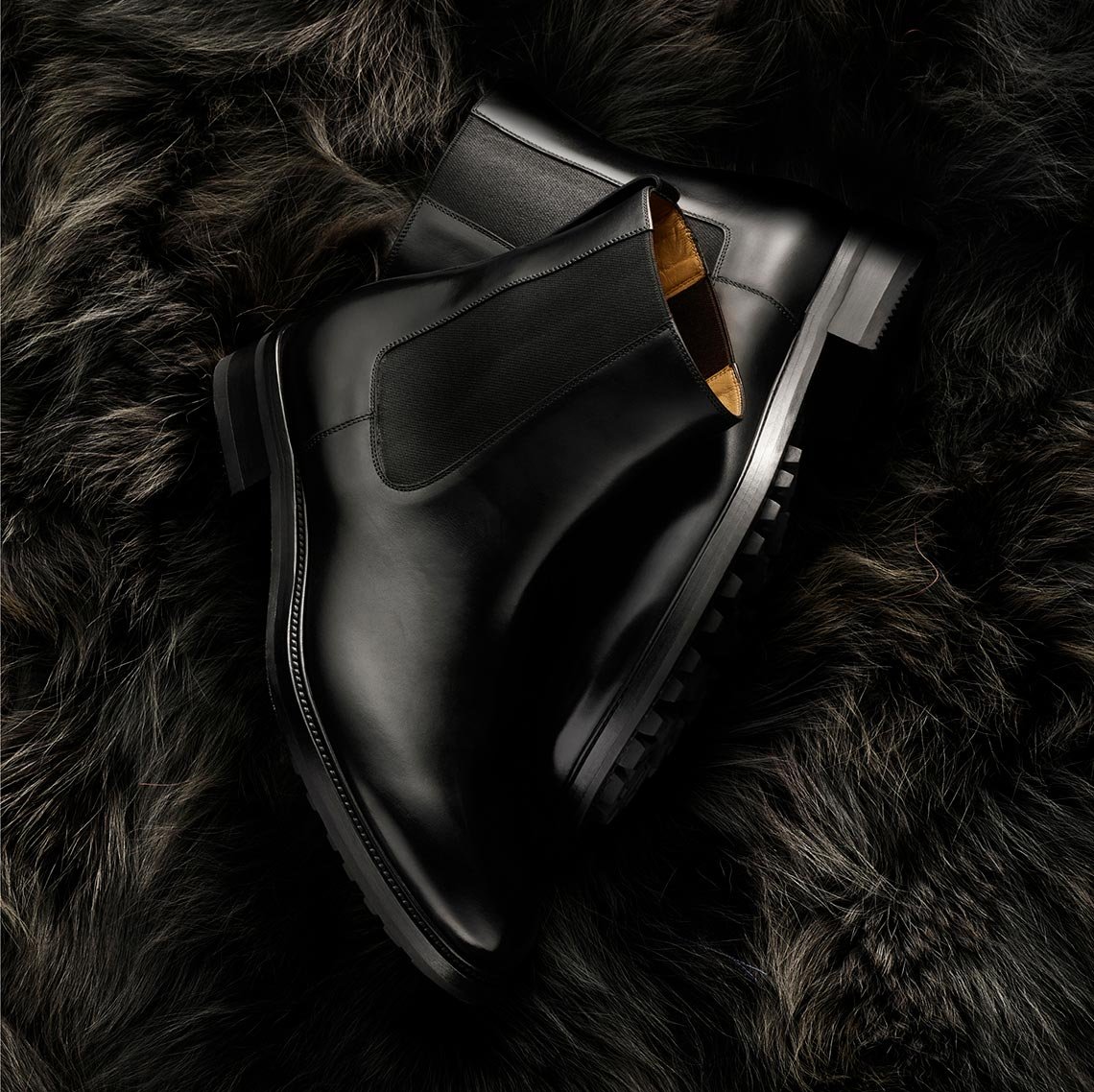 High-angle studio shot of two black leather Chelsea boots stacked diagonally against a background of soft, dark brown fur.