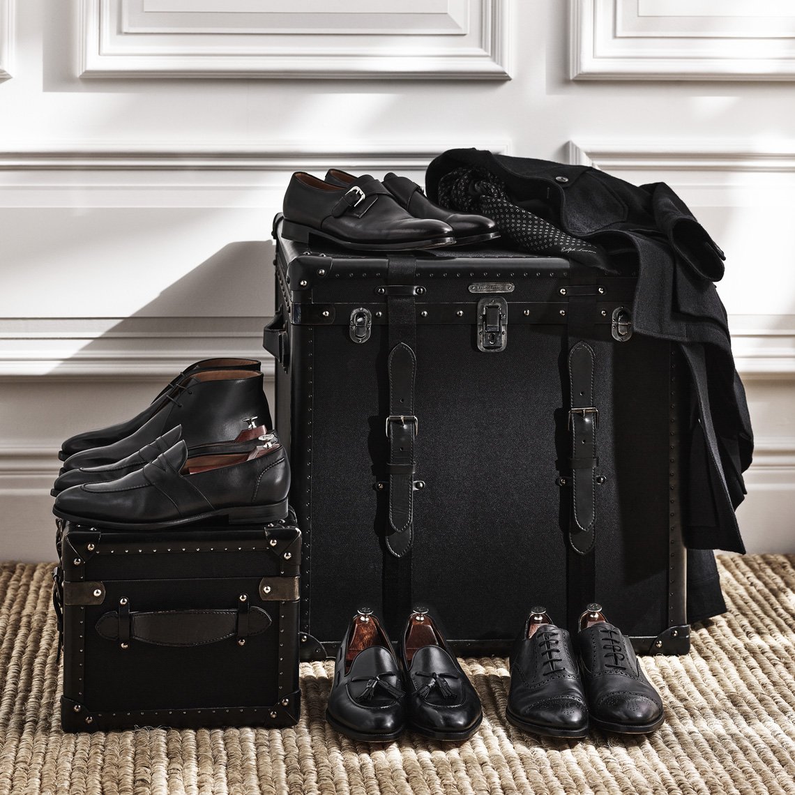 Arrangement of black leather dress shoes on stacked vintage trunks with a woven mat in front of a white paneled wall.