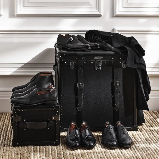 Arrangement of black leather dress shoes on stacked vintage trunks with a woven mat in front of a white paneled wall.