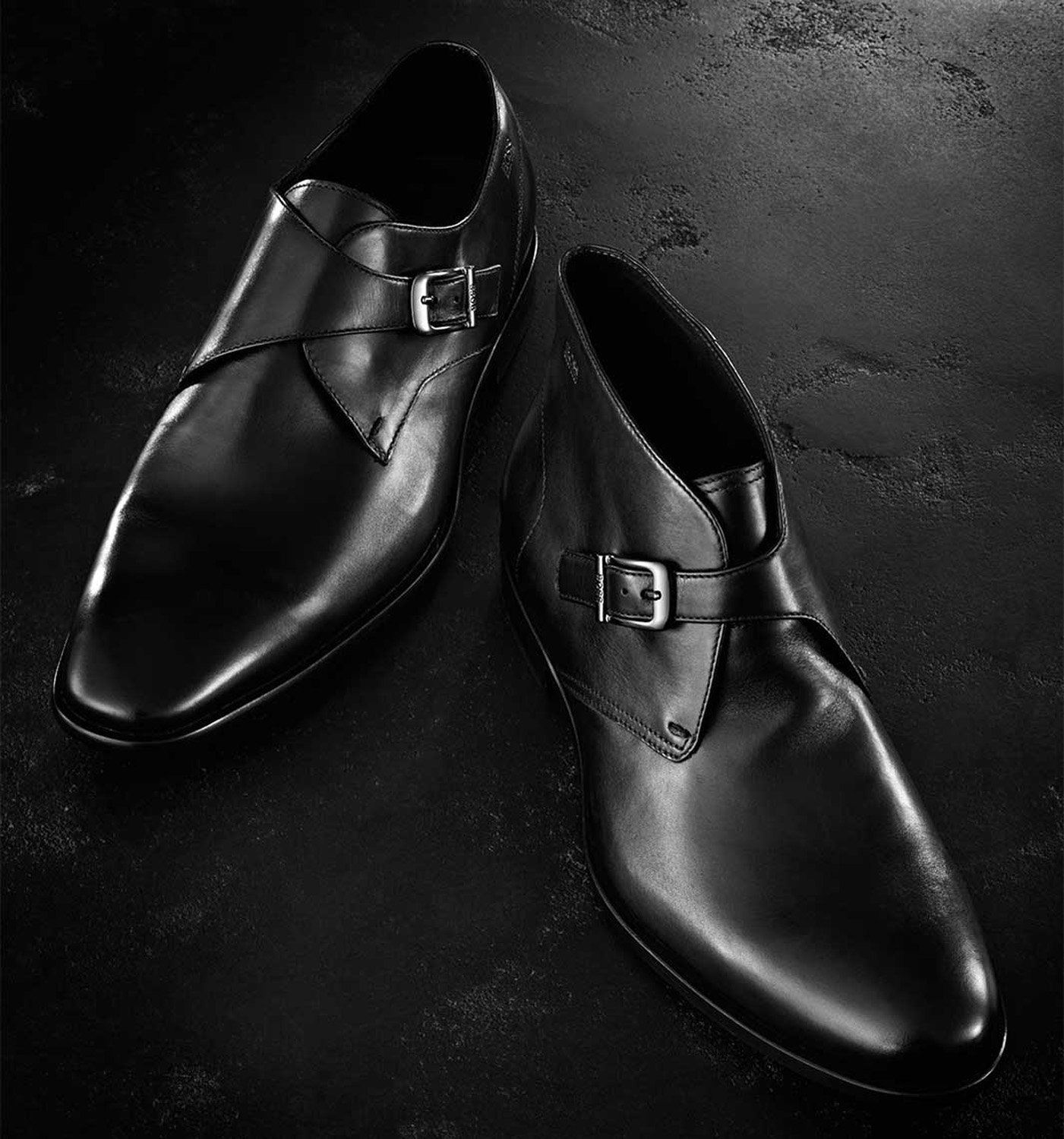 Close-up product shot of two black leather monk strap dress shoes with silver buckles, on a textured dark background, studio lighting.