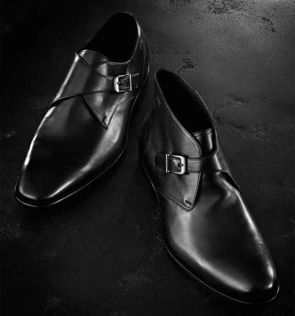 Close-up product shot of two black leather monk strap dress shoes with silver buckles, on a textured dark background, studio lighting.