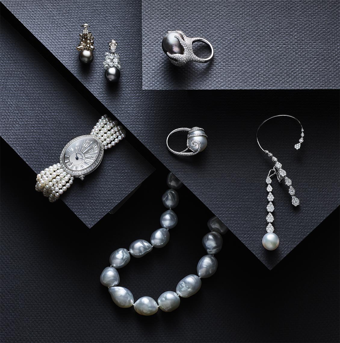 Elegant still life of pearl jewelry on a dark surface featuring a pearl watch, ring, necklace, and earrings in a geometric layout.