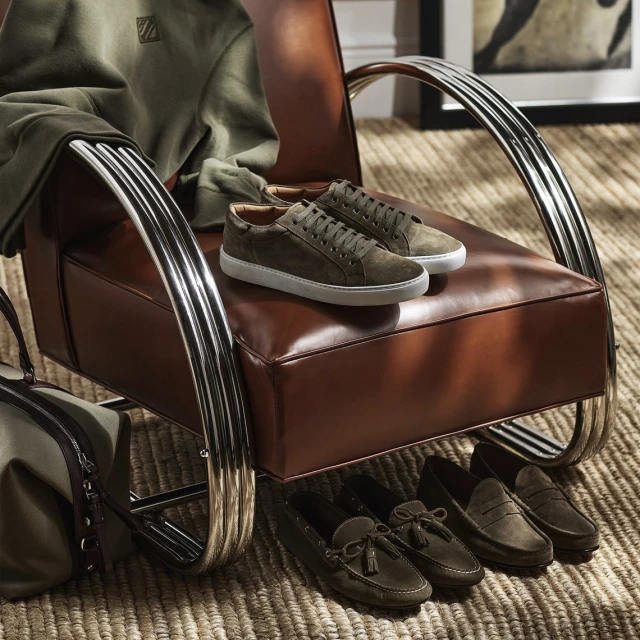 Olive green suede lace-up sneakers rest on a brown leather chair, arranged with moccasins and a duffel bag on a woven rug indoors.