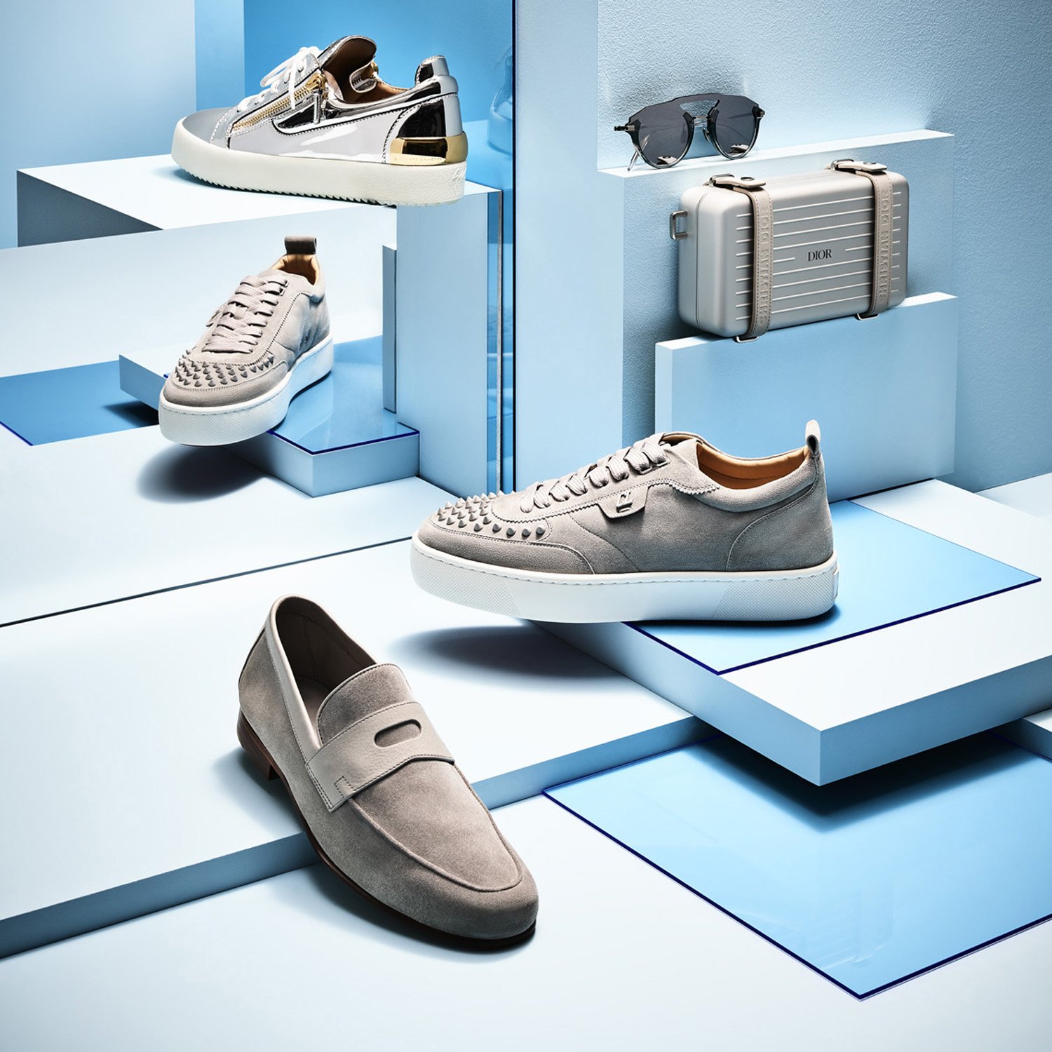 Elevated product shot showcases an assortment of luxury shoes, sunglasses, and a small suitcase on geometric platforms against a light blue background.
