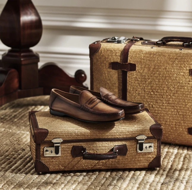 Brown leather penny loafers sit on a woven suitcase with leather trim, next to a larger suitcase and dark wood accent furniture.