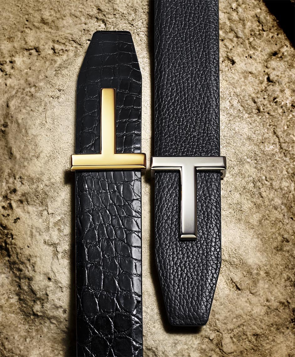Two Tom Ford belts are displayed on a textured surface, featuring 'T' logo buckles in gold and silver against dark leather straps.