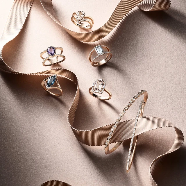Elegant rose gold rings with colorful gemstones and a diamond bracelet are displayed on a shimmering champagne-colored fabric background.