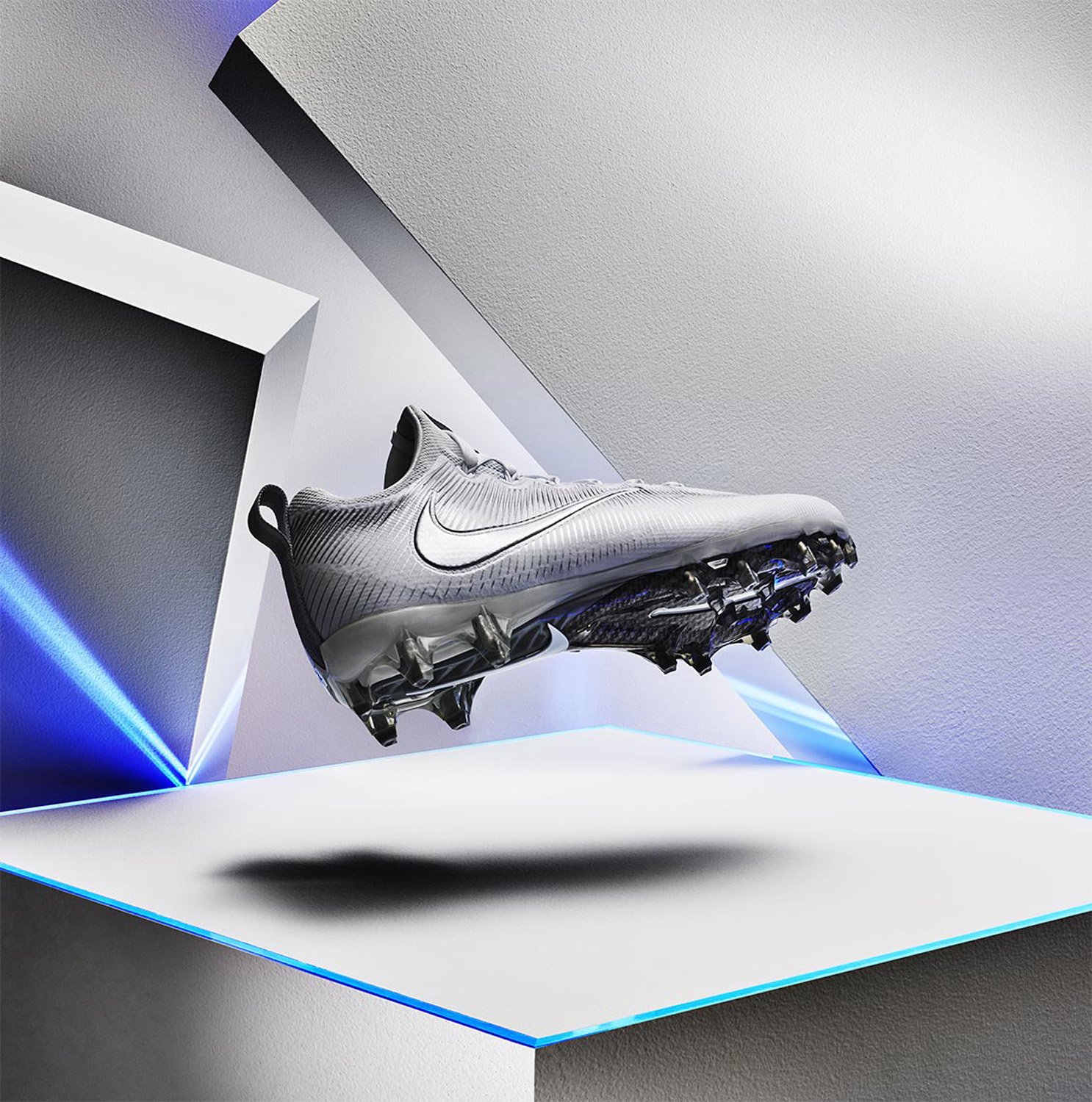 Studio shot of a silver Nike Vapor Edge Shark football cleat levitating on a white platform against a geometric background with blue lighting.