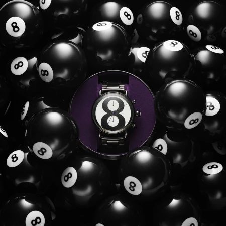 Elevated shot of a wristwatch with a black and white face displaying the number 8, displayed in the center of scattered 8-balls.