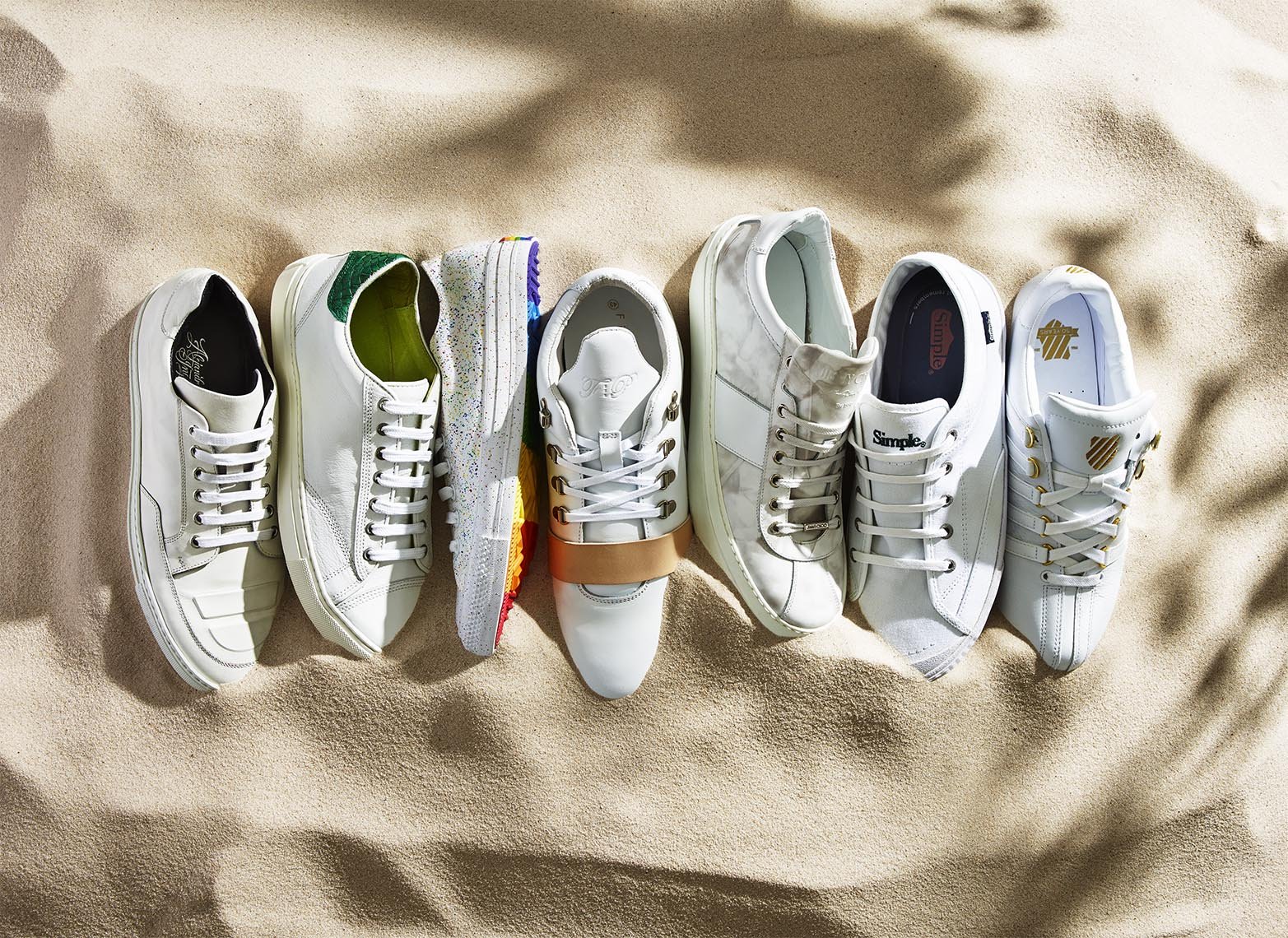 Six pairs of white sneakers are arranged on a sandy surface, showcasing different styles and brands under soft, natural lighting.