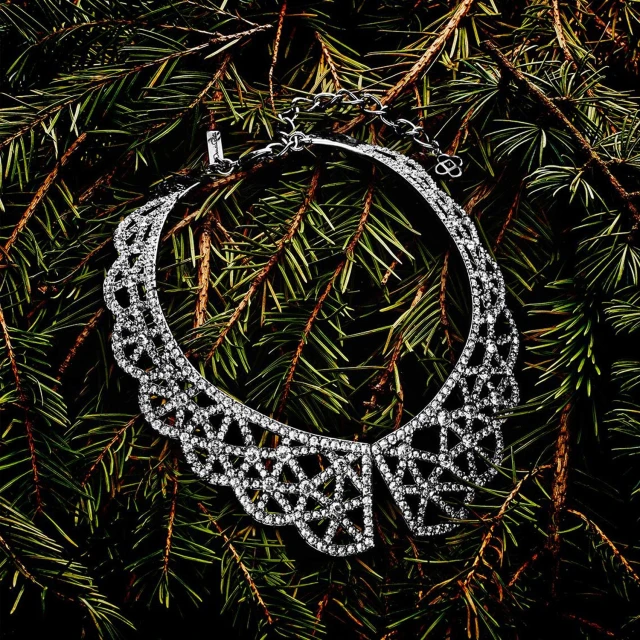Elegant silver-tone statement necklace featuring intricate geometric patterns, displayed on a bed of green pine needles against a dark background.