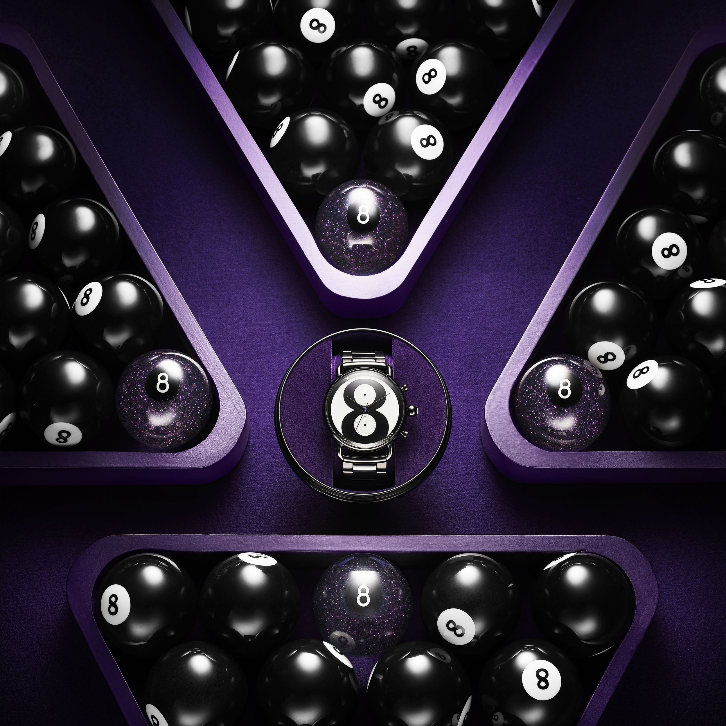 High-angle, close-up of an 8-ball watch surrounded by grouped black 8-balls in triangular wooden racks, placed on a purple surface.