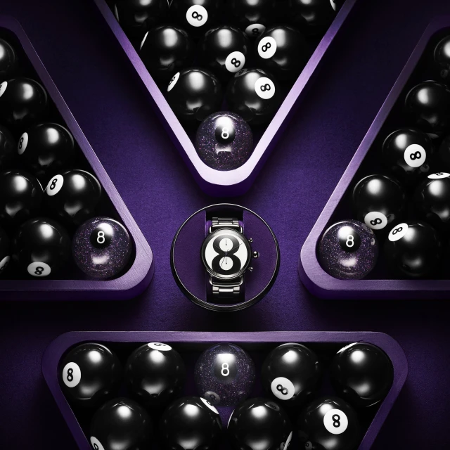 High-angle, close-up of an 8-ball watch surrounded by grouped black 8-balls in triangular wooden racks, placed on a purple surface.