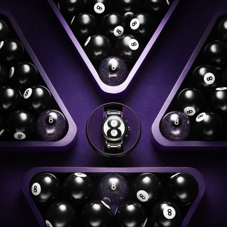 High-angle, close-up of an 8-ball watch surrounded by grouped black 8-balls in triangular wooden racks, placed on a purple surface.